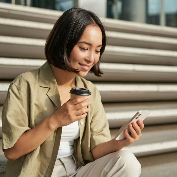 Portrait of asian woman with smartphone, drinks coffee and watches videos on mobile phone. Girl